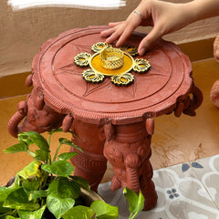 Decorative red table with gold center piece, hand reaching out, and plant on a patterned surface.