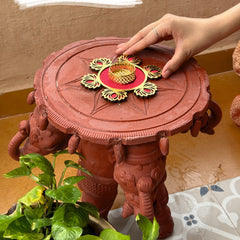 Red decorative stool with intricate design, held by a hand, on a patterned surface.
