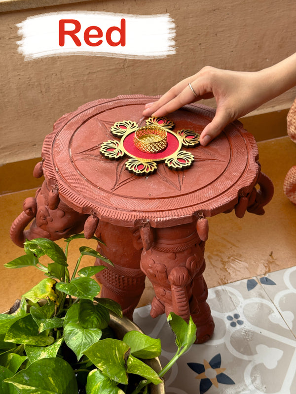 Red decorative stool with intricate design, held by a hand, on a patterned surface.