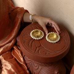 Gold earrings with pearl details on a decorative red box held by a person wearing a rust-colored garment.