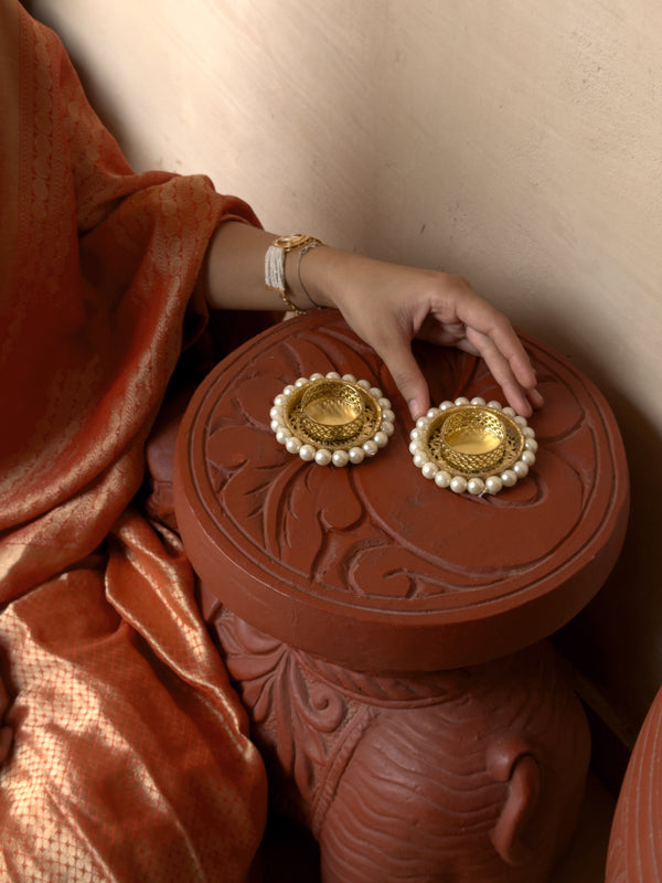 Gold earrings with pearl details on a decorative red box held by a person wearing a rust-colored garment.