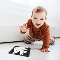 Baby in a brown outfit playing with a black card featuring a white design on a light-colored floor.