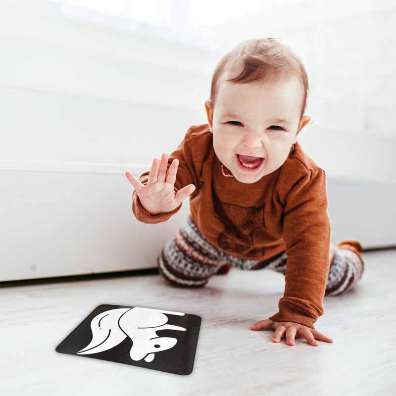 Baby in a brown outfit playing with a black card featuring a white design on a light-colored floor.
