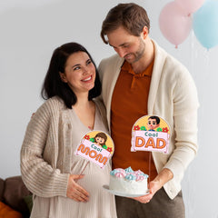 Pregnant woman and man holding a cake with 'Cool Mom' and 'Cool Dad' signs, balloons in the background.