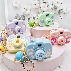 Colorful camera-shaped keychains on a white surface with flowers in the background