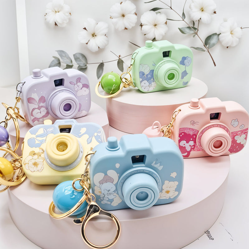 Colorful camera-shaped keychains on a white surface with flowers in the background