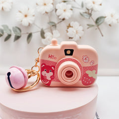 Pink camera-shaped keychain with a small bell on a white surface with flowers in the background